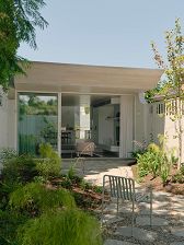 Rear facade with large open glass sliding doors and timber eave tapered to sharp edge. Lush planted garden with curved crazing paving path and terrace.