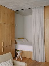 the view of a cosy bed in a studio-style apartment. A neatly made bed with write sheets and pillows is partially revealed behind an open grey curtain suspended from the ceiling. To the left we can see floor-to-ceiling wooden joinery and the corner of a grey couch, and immediately next to the bed is a small white circular stool.