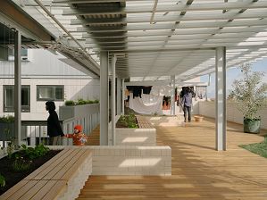 A view of the shared rooftop space which has timber decking and a semi-transparent shelter that lets the sunshine through. In the background a person is hanging washing and in the foreground and a parent is leading their toddler in an orange coat and beanie out of the shot