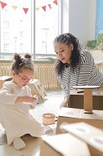 A small child and adult create buildings with cardboard in a brightly lit bedroom.