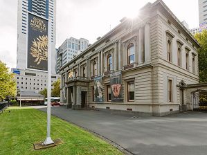 The exterior of the Former Royal Mint Building with Hellenic Museum exhibition banners on a flag pole in the foreground, and hanging from the facade.