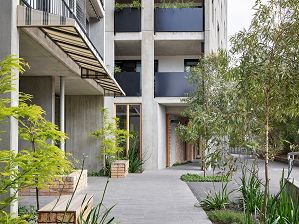 To the left is the entrance to a residential apartment building, with a cream and grey striped awning above the entrance, black planter boxes visible on the apartment balconies above, and to the right young gum trees separating the building from the concrete shared bike/pedestrian path