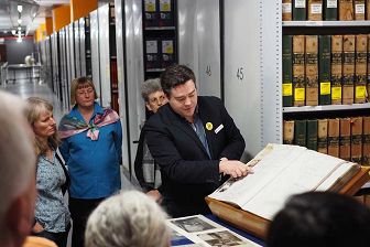 Photograph of a tour guide showing people a large historical book in the archives, surrounded by tall shelves filled with records.