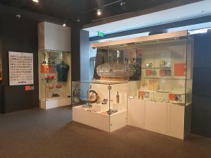 An interior image looking directly at the Interactions section of the exhibition which includes large glass display cases filled with objects, including a large commercial sized Espresso Machine from the 1950s, an early 1950s domestic past making machine, a bottle of vermouth, crockery and menus from well known Italian eateries such as The Society Restaurant, 1 1950s coloured panettone box, 1 small silver etched tea pot, 2 small bottles of olive oil,  3 large silver sports trophy one with a winged person on the top, an Australian Rules football, a well used soccer ball, a used cricket bat, a used pair of football boots, a green sports uniform on a mannequin torso with the word 