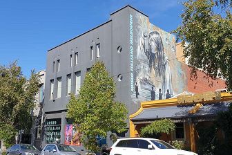 The exterior of a large two storey grey building which has a large painted colour mural by Italian multimedia artist by Alice Pasquini, covering most of the right hand-side wall of the building and the in large blue lettering are the words 'Museo Italiano Melbourne' running up the side of the wall. The mural includes an image of a torso of a woman with long dark hair looking into the distance, a ship and three black images representing people at the bottom of the mural. The large glass front window has Museo Italiano Cultural Centre stenciled in grey on the front and a brightly coloured large-scale stencilled image of an artwork by artist Matteo Volpi.