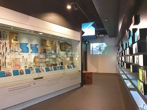 The interior image of the Settlement section of the Museo Italiano Exhibition looking toward the back wall which includes a projection of a black and white street scene in the suburb of Carlton in the early 1960s, from the Giorgio Mangiamele film, The Spag. On the left-hand side are large glass display cases filled with a wide range of objects including coffee machines, tailoring items such as scissors and ravioli rolling pin. There are 4 drawers with handles located on the bottom of the display case. On the right hand-side is a multi-coloured time-line display made up of illuminated boxes with text and images on the front each depicting a specific Australian/Italian/ World historical event such as the world wars or well-known people. There are long illuminated display cases located below the time-line boxes containing related objects, some that can be seen include books and domestic items. There is a large closed travelling box visible at the back of the space.