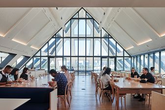 View taken in the level 6 Hub space of SEEK HQ showing people sitting and working at the tables with the large, pitched ceiling and triangular window in the background letting in plenty of natural light