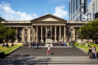 The front of State Library Victoria, with people walking up the grand steps to the building and sitting on the lawn in the sun.