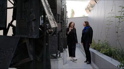 Two people are walking in conversation in an external concreted courtyard alongside a tall black metal sculpture.