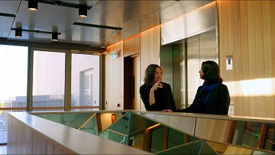 Two people are in conversation in a warmly lit room with wooden and mirrored feathers. They lean against a bannister in front of a large window with a view outside.