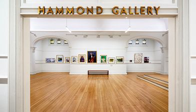 Photograph taken in the entrance to a gallery, showing the gallery sign reading 'Hammond Gallery' above. Inside is a wide space with paintings along the back wall, and two arches leading to recesses on each side.