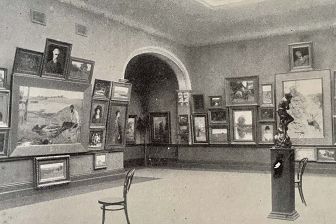 Black and white photograph of an art gallery space with the walls covered in classical paintings of various sizes. There is a sculpture of a woman on a plinth to the right, and a chair to the left in the foreground.
