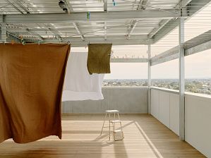 Rooftop with timber decking is sheltered by a white awning beneath which a rust coloured sheet and a khaki towel and white sheet billow in the breeze.