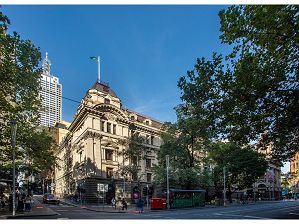 A corner image of Melbourne Town Hall's administrative building on the corner of Swanston and Little Collins Street with blue sky in the background. Pedestrians are waiting to cross the road, and post boxes and the Swanston St flower stall are visible.