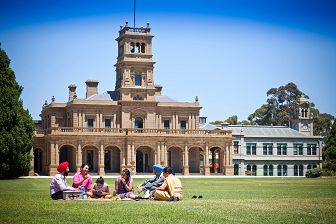 A family sits on a picnic rug on the lawn in front of the Mansion building.