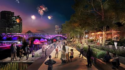 Render of people along a riverside walkway at night watching fireworks in the night sky with a bridge and high rise buildings in the background.