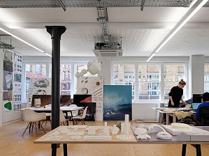 A light-filled, open plan workplace is in view with several architectural models visible on the table in the foreground. A woman wearing black with blonde hair is standing up over a computer desk, and a person in a blue jumper is working on a computer whilst sitting down. There are large art canvases pushed against the wall with large windows and design content pinned up to the left wall.