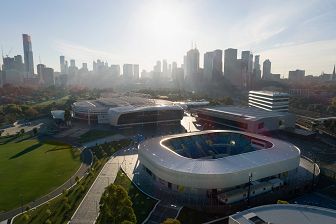 An aerial view of Melbourne Park with the City skyline in the background, the buildings are casting long shadows. It's afternoon and the sun is low. Kida Arena and CENTREPIECE are visible