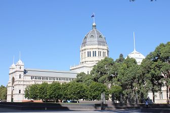A side view of the Royal Exhibition Building from Rathdowne Street, which is located in the Carlton Gardens on a clear day and under a bright blue sky. The Exhibition is the main feature of the image and is the large Italianate style nineteenth dome which is featured in the middle background and is modelled on the Dome of the Florence in Italy. There are large trees filled with green leaves in the foreground in front of which are large grey granite like modern sculptures.