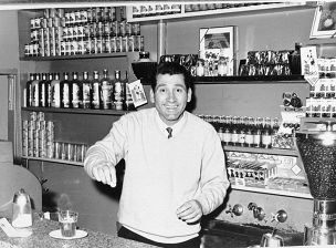 Black and white image from 1953 showing Mr Fernando Donnini well-known Carlton identity working behind the bar at the University Restaurant and Cafe at 257 Lygon Street, Carlton. Mr Donnini is wearing black pants, a white V-necked jumper on top of a white shirt and black tie. He has short dark hair, he is smiling and looking directly at the camera with both arms raised, he is holding a small round object in his right hand. There is a glass 3/4 filled with black coffee and a glass sugar container half filled with sugar on a bench in front of Mr Donnini next to a 1950s style cash register. Behind Mr Donnini there are shelves with tinned goods, bottles of soft drinks, glasses and cigarette packets. On Mr Donnini's left there is a glass container filled to the top with coffee beans. Behind Mr Donnini there is a metal sink, taps and a faucet.