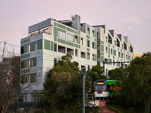 Photograph of the building with textured concrete, green timber detailing and timber windows. Bushy greenery frames the building. There is a tram going past along the adjacent tram line at speed. The dawn sky is a soft pink hue.