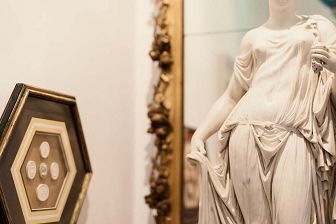On the left of this photo a hexagonal brown frame on a wall holds 5 small white circular plaster-cast coins, displayed in a '+' arrangement. On the left, a white marble statue of a woman with draped cloth over her in sits in front of a mirror.