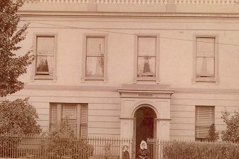 A vintage, sepia-toned, photograph of a double story single-fronted house. Two young children stand on the footpath in front of the gates of the house.