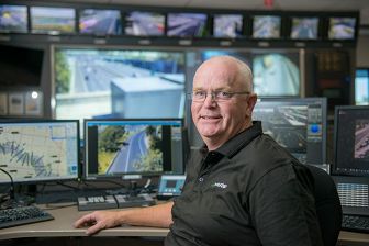 Photograph of a man facing the camera. He is sitting at a desk in the CityLink traffic control room and behind him are computer screens showing roads and traffic.