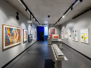 A photograph of a gallery with a concrete ceiling and grey walls. Paper-based artworks adorn the left and right walls and three artworks on plinths are situated in the middle of the room.