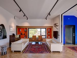 Photograph of the Justin Art House Museum apartment. The ceiling is white and the floorboards are light beige. There is a horizontal window with orange shelves on either side, holding sculptural vessels. There are two brown chairs in front of the window and two cream couches perpendicular to the chairs, facing each other with a rug and coffee table in between.