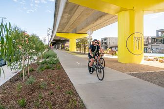 Two cyclists in black ride along the Upfield Bike Path, past bright yellow pillars supporting the elevated rail on the right and a garden bed with young gum trees to the left.