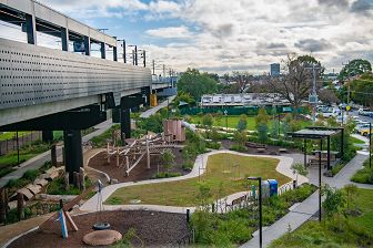 An aerial view of the Moreland Station playground. A nature playground made from wood and rope sits in the centre, with a circular swing and a slide fort on each side. A grassed area separates the playground from the barbecue and seating area. The elevated rail is heading towards Coburg, with the bike and pedestrian paths beneath.