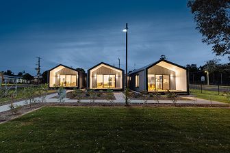 A shot of 3 houses at night. They are lined up in a row with pitched roofs. The interior lights of the homes are on, while the outside looks like dusk.
