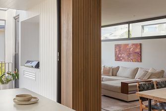 The interior of a home, to the left you can see the outdoor patio and barbeque, to the right looks through to the lounge where there's a cream sofa. The colours are light, and the wall separating the two spaces is wood.