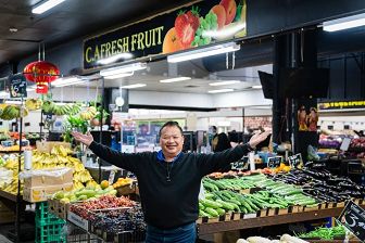 A man stands in front a fruit stand with his arms in an open embrace. Above him, a sign reads CA Fresh Fruits.