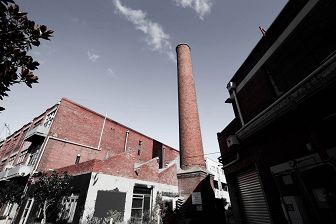Image looks up at a red brick chimney flanked by two red industrial brick buildings.