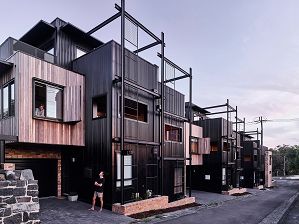 Photo of a series of homes clad in timber and black steel with brick elements. The man is in shorts looking at an upstairs window where a woman is leaning out of a window.