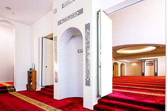 Interior of the Albanian Mosque of the prayer area. There is burgundy coloured carpet with gold lines which indicate where people stand to pray. There is a close up of the mihrab, which is the niche where the Imam leads the prayer and indicating the direction to Mecca.