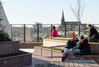 A woman in a pink top sits in the distance and a group of three students sit on planter steps on the terrace during a break on a sunny winter’s day, with views to a large church