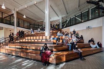 students sit on the timber tiered seating within the atrium. Another group of students walk up the stairs of the atrium which are integrated with the seating