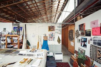 Interior colour photograph of an artist studio below an exposed timber structured roof. Artworks adorn the walls and the table in the foreground.