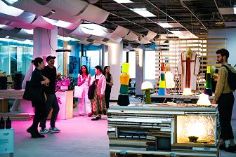 A colour photograph of a retail space made from waste materials lit up with a pink light. There are people moving through the space looking at the various art objects situated on top of plinths and tables.
