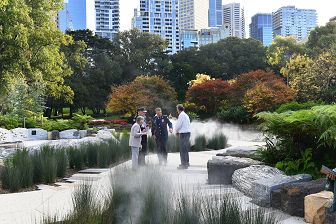 A woman with three men stand on a stone path within the Memorial. There are clouds of mist from a fountain. The mist floats above green reed beds. On the right are green ferns. Behind the memorial are mature trees that have changed colour in autumn. In the background is the Melbourne cityscape.