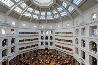 (1) Domed, high-ceilinged white room with skylights in roof. Lower down there are 6 bookshelf balconies spanning 3 levels filled with books. Below that are wooden bookshelves and desks on the ground floor, full of people. (2) Outside shot of State Library Victoria, grand building with columns at entrance. Sunny day. People walking across the forecourt. (3) Many small, wooden drawers, with handwritten labels and stickers on front of each. Some are pulled out, and showing stacks of cards, and others are not. (4) Alternative entrance of State Library Victoria, with an indicator that it is entrance 6. Also has columns and is quite grand, warm lights are on inside.