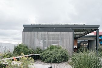 a cloudy day on a rooftop terrace with timber flooring and planted native shrubs growing around a wooden bench seat, behind which a portion of the rooftop is protected by a steel framed shelter with wooden cladding on the sides and solar panels visible mounted on top.
