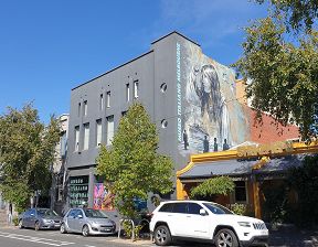 The exterior of a large two storey grey building which has a large painted colour mural by Italian multimedia artist by Alice Pasquini, covering most of the right hand-side wall of the building and the in large blue lettering are the words 