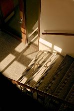 View from above down a staircase, light from the left casts shadows of the bannister across the floor and stairs.
