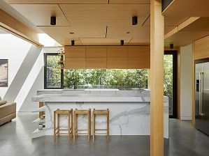 The kitchen of the Timber-Lantern House, with timber clad elements, a sculptural white island bench and feature glazed window looking out to greenery.