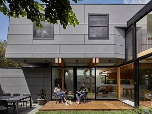 A two-storey home addition clad in grey concrete panels, showing a view to the living room beyond with home-owners relaxing on the deck with their cute white dog