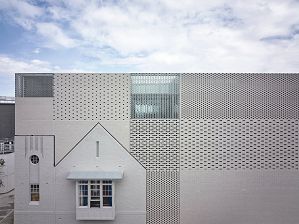 Facade of the Melbourne Holocaust Museum, a four-story rectangular building built of grey bricks alternating with glass bricks. The left-hand-side of the building includes the facade of the smaller two-story original museum built in 1984, which has a bay window and pointed roof.